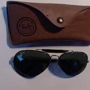 May include: Aviator sunglasses with dark lenses and a silver frame, resting next to a brown leather case. The case has a snap closure and a Ray-Ban logo. The sunglasses have a classic, timeless design.