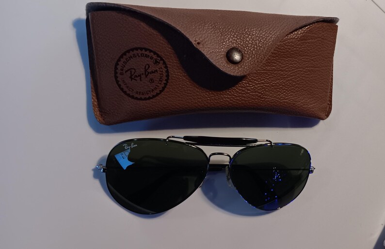 May include: Aviator sunglasses with dark lenses and a silver frame, resting next to a brown leather case. The case has a snap closure and a Ray-Ban logo. The sunglasses have a classic, timeless design.