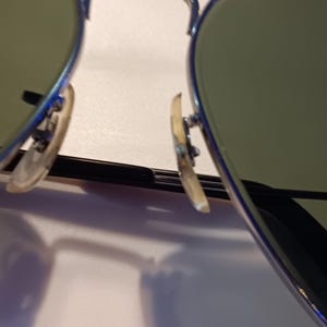 May include: Close-up of aviator sunglasses with a silver frame and green-tinted lenses. The nose pads are a light tan color. The lenses reflect a light source.