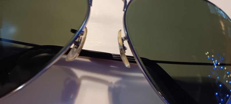 May include: Close-up of aviator sunglasses with a silver frame and green-tinted lenses. The nose pads are a light tan color. The lenses reflect a light source.