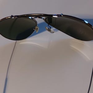 May include: Sunglasses with a silver frame and dark lenses. The sunglasses have a classic aviator style with a double bridge and thin, black temples. The lenses are slightly reflective.