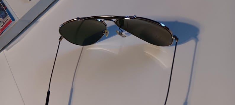 May include: Sunglasses with a silver frame and dark lenses. The sunglasses have a classic aviator style with a double bridge and thin, black temples. The lenses are slightly reflective.
