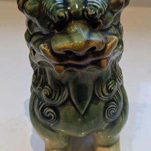 May include: A green ceramic foo dog figurine with detailed facial features and a curled mane. The figurine has a glazed finish and stands on four legs.