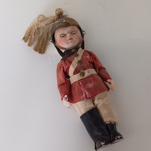 May include: A vintage doll dressed as a soldier, wearing a red and white uniform with a gold helmet and a white plume. The doll has a painted face with closed eyes and is lying down.
