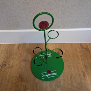 Pode incluir: Porta-garrafas de metal verde com an&eacute;is de borracha preta para segurar garrafas. O porta-garrafas tem uma base redonda com o logotipo Tanqueray e o texto "EST. LONDON 1830".