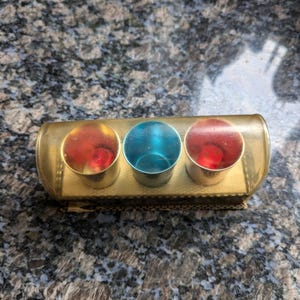 Puede incluir: Un recipiente rectangular de plástico transparente de color dorado con tres pequeñas tazas redondas de colores en su interior. Las tazas son rojas, azules y rojas. El recipiente está sobre una superficie gris moteada.