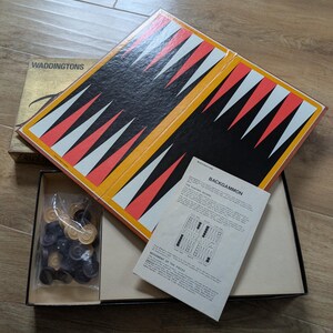 May include: A vintage backgammon game set in a box. The game board is black and white with red and yellow accents. The box includes the game board, dice, and counters.
