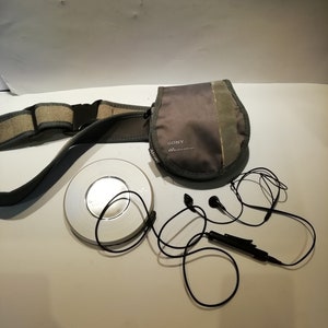 Könnte beinhalten: Ein silberner Sony Walkman CD-Player mit einer grauen Tragetasche und schwarzen Kopfhörern. Der Player befindet sich auf einer weißen Oberfläche.