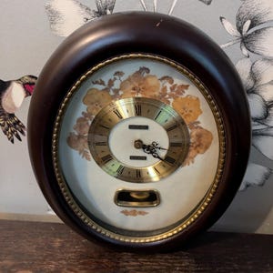 May include: A vintage oval clock with a dark wood frame and a gold trim. The clock face is white with gold Roman numerals and a gold second hand. The clock is decorated with pressed dried flowers.