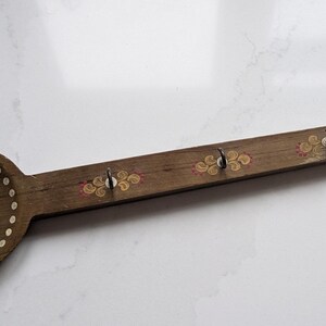 May include: Wooden spoon-shaped wall hook with a dark brown finish. The spoon bowl features a floral design with white and yellow accents. The handle has five metal hooks and is decorated with floral patterns.