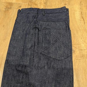 May include: Dark blue denim jeans, folded and viewed from the back. The jeans feature a single back pocket with a decorative stitch pattern and a visible seam running down the center.