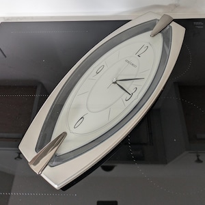 Puede incluir: Un reloj de pared Seiko plateado, de forma ovalada, con esfera blanca. El reloj presenta manecillas y números negros, con el nombre de la marca Seiko impreso en la parte superior. Diseño moderno con detalles plateados en la parte superior e inferior.