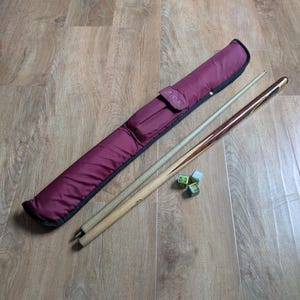 Op de afbeelding: Een bordeauxrode biljartkeu-etui met zwarte rand, twee biljartkeuen en drie groenblauwe krijtwürfel. Eén keu is lichtgekleurd hout, de andere is donker hout met gele accenten. De etui heeft de tekst "B.C.E."