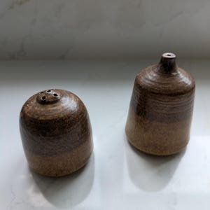 May include: A pair of handcrafted ceramic salt and pepper shakers. The salt shaker is a rounded shape with three holes on top. The pepper shaker is a taller, bell-shaped design. Both are brown with a darker band around the middle.