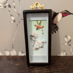 Op de afbeelding: Een ingelijst kunstwerk met een vogel en bloemen gemaakt van veren. De lijst is zwart met een gouden bovenkant. Het kunstwerk heeft Chinese karakters in de rechterbenedenhoek.