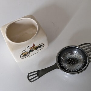 Op de afbeelding: Een witte theepot van keramiek met een vintage auto-ontwerp en de tekst "Earl's Automobile" aan de zijkant. Een zilveren theezakje met een handvat en een gaasbodem is ook afgebeeld.