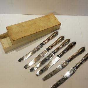 May include: Set of six silver butter knives with ornate, twisted gold-colored handles. The blades are silver and slightly serrated. The knives are arranged next to an open, aged cardboard box.