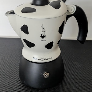 Puede incluir: Una cafetera espresso Bialetti Mukka Express blanca y negra. La cafetera tiene un patrón de vaca con manchas negras. El asa y la parte superior son negras. El nombre y el logotipo de la marca son visibles.