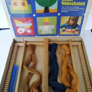 May include: A wooden loom with a blue comb, brown, blue, and yellow yarn, and a box with images of a house, tree, butterfly, and person. The box reads "Schul-Webrahmen" and "Webbreite 25cm".