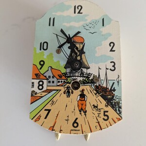May include: A vintage wooden clock with a colourful illustration of a windmill, houses, and boats. The clock face has black numbers and hands, and the clock is labelled "Germany".