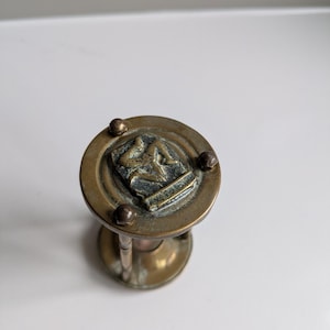 Op de afbeelding: Een vintage messing zandloper met een gedetailleerd embleem aan de bovenkant. De zandloper heeft een cirkelvormige boven- en onderkant, verbonden door slanke steunen. Het embleem heeft een gedetailleerd ontwerp en het messing heeft een licht verouderde patina.