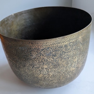May include: A round brass bowl featuring an intricate, dark-etched floral and foliate pattern covering the exterior surface. The design includes repeating scrollwork and stylised figures, with a decorative border near the rim and a smooth interior.