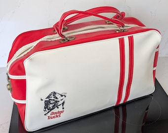 Pode incluir: Uma mala de viagem vintage vermelha e branca com alças e detalhes vermelhos. A mala apresenta duas listras vermelhas verticais e um gráfico em preto e branco de um touro com o texto "Dodge Trucks".