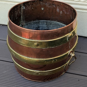 May include: A copper-colored barrel-shaped container with brass bands and a brass handle. The container has a dark interior and is sitting on a wooden surface. The bands are secured with rivets. The container is likely used for decorative purposes.