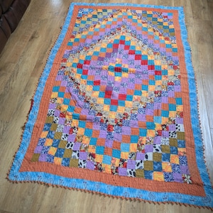 May include: A colorful patchwork quilt with a vibrant design. The quilt features a central diamond pattern surrounded by squares of various colors, including red, orange, blue, and purple. The edges are trimmed with a blue border and a ruffled edge.