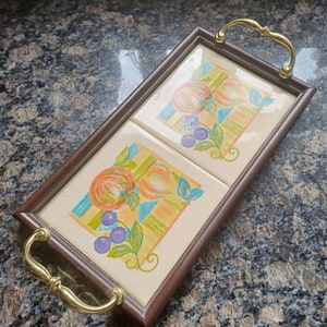 May include: A rectangular serving tray with a brown wooden frame and two cream-colored tile inserts. Each tile features a colorful illustration of fruit, including oranges, grapes, and leaves. The tray has gold-colored handles.