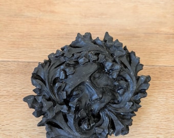 Può includere: Una scultura nera, intagliata in modo intricato, di un volto di Green Man, circondato da un fogliame dettagliato. La scultura è realizzata in un materiale scuro e poggia su una superficie di legno chiaro. Il volto è centrato nel disegno foglioso.