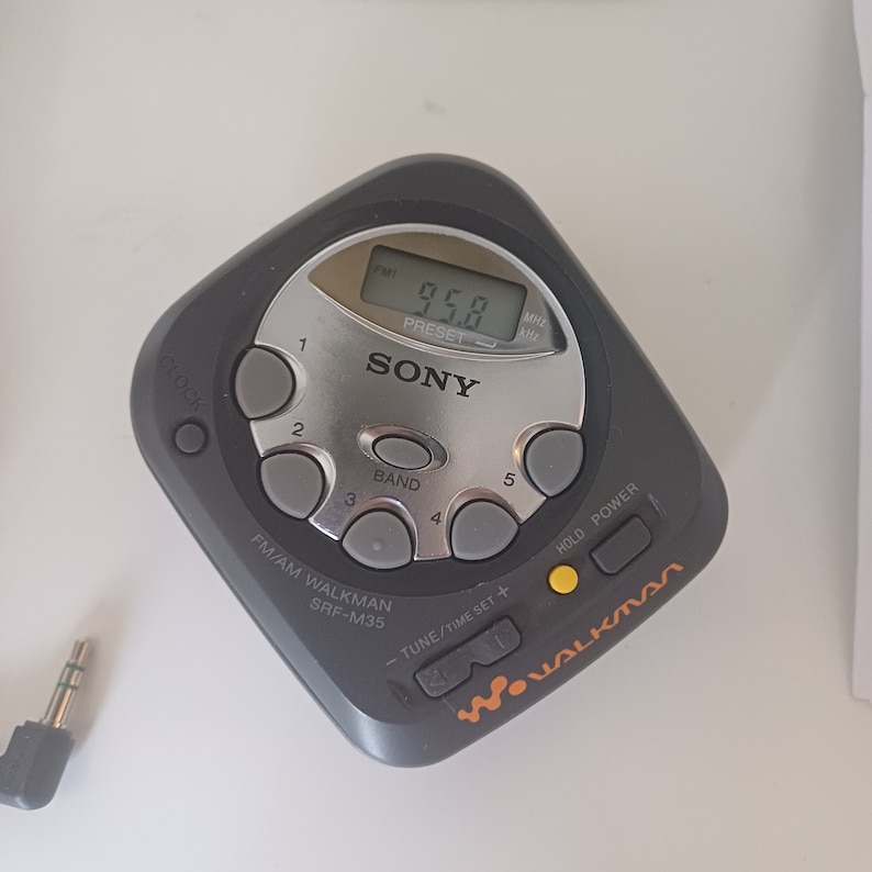 May include: A black Sony Walkman SRF-M35 AM/FM radio with a silver faceplate and a digital display showing 95.8. The radio has a number of buttons, including a power button, a tune/time set button, a hold button, and a band button. The radio also has a clock button and a preset button.