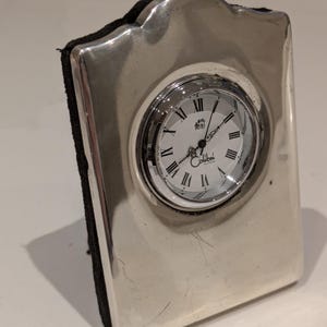 May include: A silver desk clock with a rectangular frame and a round clock face. The clock face features Roman numerals and the brand name "Colibri". The back of the clock is black.
