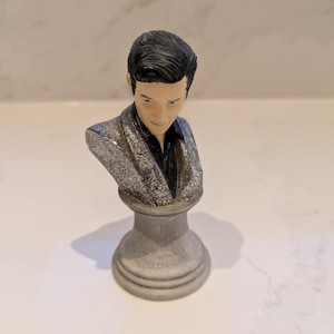 May include: A small bust of a man with dark hair and blue eyes, wearing a black shirt and a silver sequined jacket. The bust is mounted on a grey pedestal. The sculpture is a decorative item.