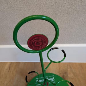 Pode incluir: Porta-copos de metal verde com um logotipo vermelho e branco que diz "TAMBERELLI'S FINE FOOD AND DRINK". O porta-copos tem quatro ganchos para copos e uma base redonda.