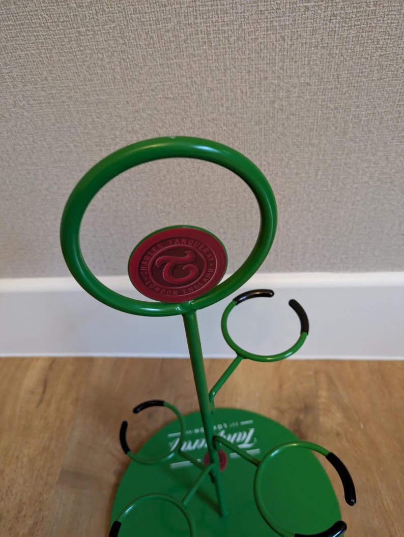 Pode incluir: Porta-copos de metal verde com um logotipo vermelho e branco que diz "TAMBERELLI'S FINE FOOD AND DRINK". O porta-copos tem quatro ganchos para copos e uma base redonda.