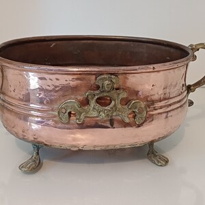 Pode incluir: Um vaso oval de cobre com três pés e duas alças de latão ornamentadas. O vaso tem um acabamento martelado e uma banda de latão em torno da parte superior.