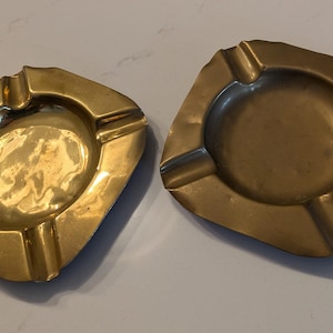 Op de afbeelding: Twee vintage messing asbakken. Elke asbak heeft een vierkante vorm met afgeronde hoeken en een cirkelvormige inkeping in het midden. De asbakken hebben een gouden kleur en zijn voorzien van vier sigarettensteunen.