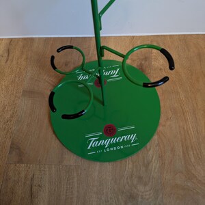 Pode incluir: Porta-copos de metal verde com quatro porta-copos com borda preta e uma base circular com o texto "Tanqueray. Est. London 1830."