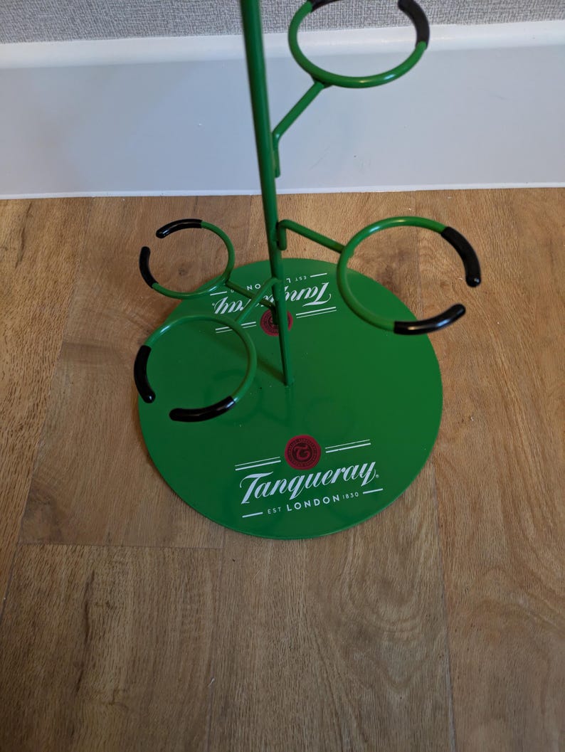 Pode incluir: Porta-copos de metal verde com quatro porta-copos com borda preta e uma base circular com o texto "Tanqueray. Est. London 1830."
