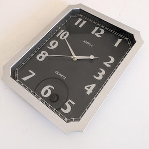 May include: A silver framed black clock with white numbers and hands. The clock face says "AMSUEA QUARTZ".