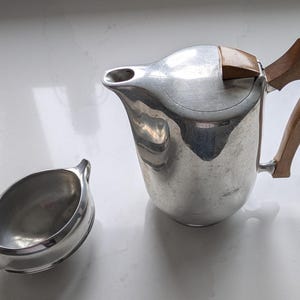 Puede incluir: Una tetera de plata con asa y tapa de madera, y una jarra de crema de plata a juego.