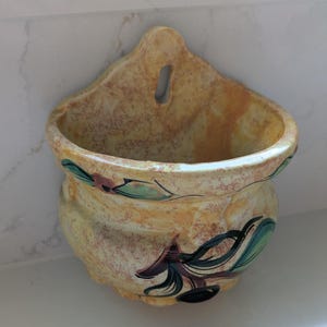 Puede incluir: Un macetero de pared vintage con un acabado moteado amarillo y marrón. El macetero de cerámica presenta un diseño floral decorativo en tonos verdes, morados y rosas. Tiene un cuerpo redondeado y un ojal para colgar.