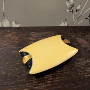 May include: An Art Deco brooch with a cream-coloured, shield-shaped top and black and gold detailing. The brooch is set against a dark wooden surface.