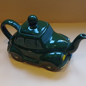 Puede incluir: Una tetera de cerámica verde con forma de coche antiguo. La tetera tiene un pico y un asa, y un detalle amarillo en la parte delantera.