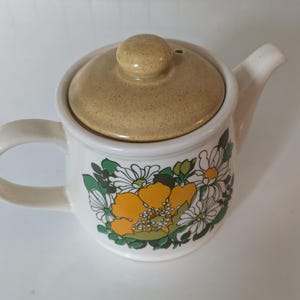 以下が含まれることがあります： 花柄の白い陶器製のティーポット。ティーポットは、ノブと注ぎ口が付いた蓋が付いています。花柄は、黄色、白、緑の花で構成されています。
