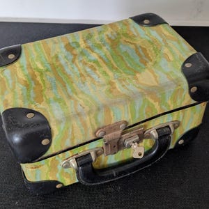 May include: A small vintage suitcase with a green, yellow, and blue marbled pattern. The corners are reinforced with black metal, and it has a black handle and a metal clasp with a key.