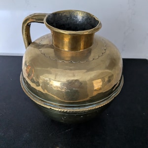 Pode incluir: Um jarro redondo de latão com gargalo largo, uma única alça e uma faixa decorativa recortada ao redor da base do gargalo. O corpo apresenta uma costura horizontal com uma textura de corda trançada que circunda o meio do recipiente.