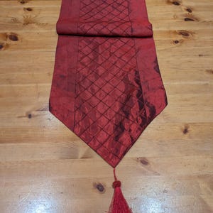 Puede incluir: Un camino de mesa rojo oscuro con un patrón de diamantes y una borla. El camino de mesa está hecho de una tela brillante y está colocado sobre una mesa de madera. La borla está al final del camino de mesa.