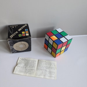Puede incluir: Un cubo de Rubik con una caja negra con la etiqueta "CHALLENGE PUZZLE" y un pequeño folleto de instrucciones. El cubo tiene cuadrados de colores en rojo, azul, verde, naranja, amarillo y blanco.